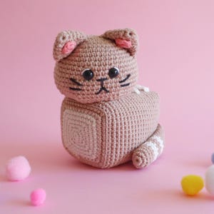 Puede incluir: Un juguete de gato tejido a ganchillo en tonos beige y blanco, con detalles rosas en las orejas. El gato tiene ojos y bigotes negros. La cola tiene rayas blancas. Varias bolas de pelo de colores están esparcidas.