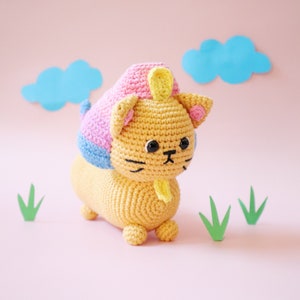 May include: A crocheted lion toy with a pink, blue, and yellow mane. The lion has black eyes and a yellow tongue sticking out. The toy is sitting on a pink surface with green paper grass.
