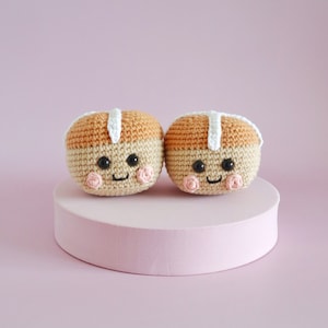 May include: Two crocheted hot cross buns with smiling faces. The buns are light brown with white icing and pink cheeks. They are sitting on a pink platform.
