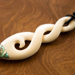 Pode incluir: Um pingente de osso branco com um design em espiral e uma incrustação de madrepérola. O pingente está em um cordão preto.