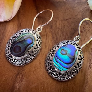 Peut inclure: Une paire de boucles d'oreilles pendantes de forme ovale avec des incrustations de nacre d'ormeau irisée serties dans des montures en métal argenté oxydé. Chaque boucle est ornée d'un motif floral répétitif autour de la nacre et d'un crochet incurvé.