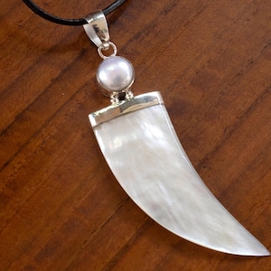 May include: A pendant necklace with a large, pearly white, curved tooth-shaped pendant. The pendant is attached to a silver-coloured bail and a small, round pearl. The necklace has a black cord.