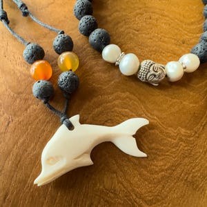 Könnte beinhalten: Ein weißer, geschnitzter Delfin-Anhänger an einer Halskette aus schwarzen Lavasteinen und orangefarbenen Perlen. Die Halskette enthält auch einen silbernen Buddha-Anhänger und weiße Perlen.