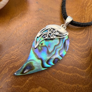 Op de afbeelding: Een druppelvormige hanger van abalone-schelp met iriserende blauwe en groene tinten. De bovenkant van de schelp is afgewerkt met een zilverkleurig metalen onderdeel met gedetailleerd krulwerk. De hanger hangt aan een zwart koord.