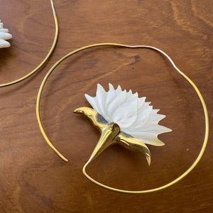 Peut inclure: Une paire de boucles d'oreilles créoles avec un grand élément floral blanc sculpté fixé à un fil métallique doré fin. La fleur présente des pétales pointus superposés et est fixée à l'anneau circulaire par une base en métal doré poli.