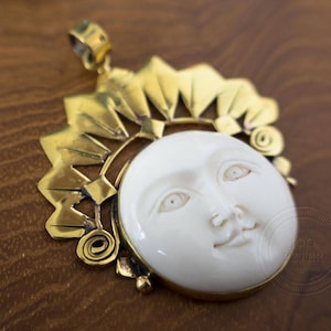 Peut inclure: Pendentif doré représentant un visage de soleil blanc sculpté. Le visage du soleil est entouré d'un cadre doré décoratif avec des rayons de soleil et des spirales. Le pendentif est probablement en laiton ou métal similaire.