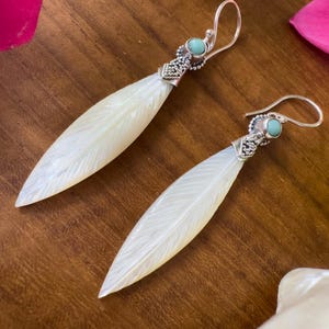 Può includere: Un paio di orecchini pendenti con design a forma di piuma. Gli orecchini hanno una parte superiore in metallo color argento con una piccola pietra turchese. Il corpo a forma di piuma è bianco con venature dettagliate. Presentati su una superficie di legno.