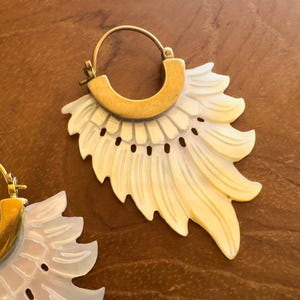 Könnte beinhalten: Nahaufnahme von zwei goldenen Creolen mit einem einzigartigen Design. Jede Ohrring hat einen goldenen Reifen und einen cremefarbenen, fächerförmigen Anhänger mit gewelltem Rand und kleinen schwarzen Punkten. Die Ohrringe sind vor einem braunen Hintergrund platziert.
