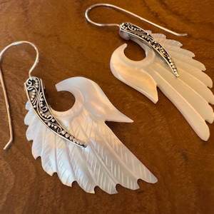 Op de afbeelding: Een paar oorbellen met een vleugelontwerp. De vleugels zijn wit met gedetailleerde, veerachtige patronen. Elke oorbel heeft een zilverkleurig decoratief accent en een zilveren haak. De oorbellen worden gepresenteerd op een bruin houten oppervlak.