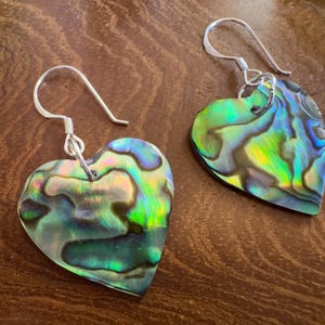 Peut inclure: Une paire de boucles d'oreilles en forme de cœur avec des coquillages d'ormeau irisés. Les boucles d'oreilles ont des crochets argentés. Les coquillages d'ormeau affichent un mélange de teintes vertes, bleues et jaunes. Les boucles d'oreilles sont présentées sur une surface en bois.