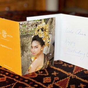 May include: A greeting card with a photo of a woman in traditional Balinese attire. The card is open, revealing the photo and handwritten text that reads "Liebe Anna, Happy Birthday". The card has a yellow cover with the text "Bali Moon".