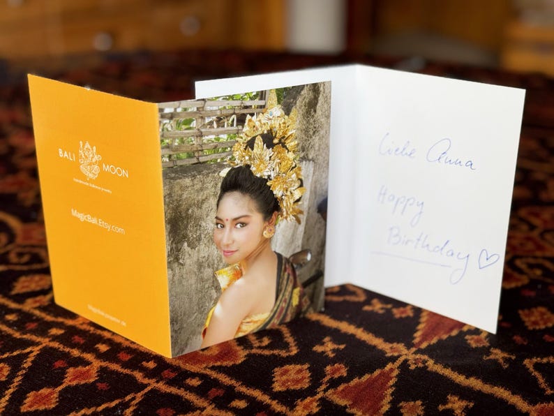 May include: A greeting card with a photo of a woman in traditional Balinese attire. The card is open, revealing the photo and handwritten text that reads "Liebe Anna, Happy Birthday". The card has a yellow cover with the text "Bali Moon".