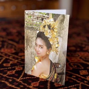 May include: A folded greeting card featuring a woman in traditional Balinese attire. She wears a gold headdress and earrings, with a patterned shawl. The background shows a stone wall and foliage. The card is on a patterned surface.