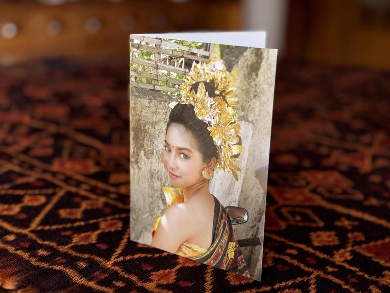 May include: A folded greeting card featuring a woman in traditional Balinese attire. She wears a gold headdress and earrings, with a patterned shawl. The background shows a stone wall and foliage. The card is on a patterned surface.