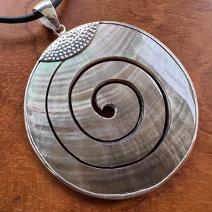 Könnte beinhalten: Ein großer, runder Anhänger mit einem Spiraldesign. Der Anhänger hat einen Perlmutt-Mittelteil mit einem silberfarbenen Metallrahmen und einer Öse. Er hängt an einem schwarzen Band.