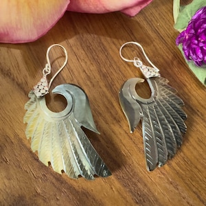 May include: A pair of silver-toned earrings with a wing design. The earrings have a pearlescent, iridescent finish with a feather-like texture. The earrings are displayed on a wooden surface with flower petals.