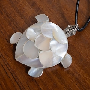 Pode incluir: Um colar com um pingente de tartaruga de prata com incrustações de madrepérola branca. O colar está em um cordão preto.