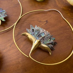 May include: A pair of large hoop earrings featuring a thin gold-toned metal wire frame. Each earring is adorned with a decorative element shaped like a flower, crafted from iridescent abalone shell with a polished gold-toned metal base.