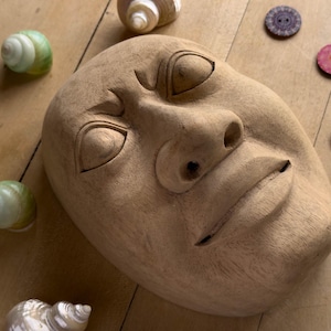 Könnte beinhalten: Eine hellbraune Holzmaske mit neutralem Gesichtsausdruck, geschnitzt mit mandelförmigen Augen, einer markanten Nase und einem geraden Mund. Die Maske ist von mehreren kleinen, irisierenden spiralförmigen Muscheln und bunten Knöpfen umgeben.
