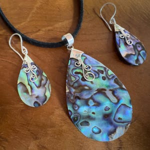 Könnte beinhalten: Schmuckset aus Abalone-Muscheln mit einer Anhänger-Halskette und passenden Ohrringen. Die tropfenförmigen Stücke zeigen irisierende Blau-, Grün- und Lilatöne. Akzente aus silberfarbenem Metall und eine schwarze Kordel vervollständigen die Halskette.