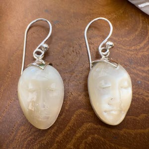 Pode incluir: Um par de brincos pendentes com rostos esculpidos em madrepérola com olhos fechados. Cada peça de concha oval é fixada numa tampa de metal prateado polido e ligada a um gancho de fio prateado para orelhas furadas.