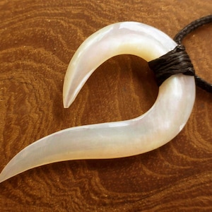 Op de afbeelding: Een parelmoer, gebogen hangerketting die lijkt op een vishaak. De hanger is bevestigd aan een zwart koord. De ketting staat tegen een bruine achtergrond met houtnerf. De hanger is gemaakt van een lichtgekleurd, iriserend materiaal.