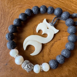 Może przedstawiać: Bransoletka z czarnego wulkanicznego kamienia z białymi perłami i białą rzeźbą z kości przedstawiającą stylizowaną figurę. Bransoletka jest związana czarną sznurówką.