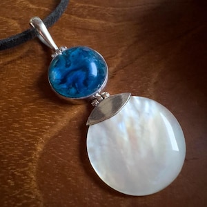Könnte beinhalten: Eine Anhängerkette mit einer runden, irisierenden Perlmutt-Scheibe und einem kleineren, runden blauen Stein mit wirbelnden Mustern. Der Anhänger ist durch ein silberfarbenes Metallstück verbunden und hängt an einer schwarzen Kordel.
