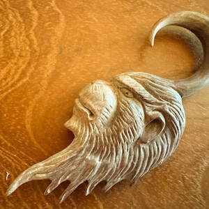 Könnte beinhalten: Eine geschnitzte Holzskulptur eines Fabelwesenkopfes mit einem langen, gebogenen Horn und detaillierten Gesichtszügen. Die Skulptur ist hellbraun und liegt auf einer Holzoberfläche.