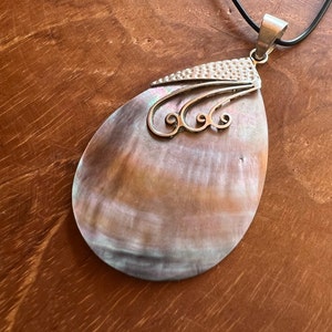 Puede incluir: Un collar con colgante en forma de lágrima con una concha de nácar. El colgante tiene una parte superior de metal plateado con un diseño texturizado y remolinos decorativos. El collar cuelga de un cordón negro.