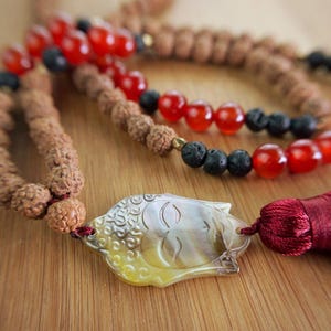 Könnte beinhalten: Nahaufnahme einer Mala-Perlenkette mit roten, schwarzen und braunen Perlen. Die Halskette hat einen geschnitzten Buddha-Anhänger und eine rote Quaste. Die Perlen sind auf einer braunen Schnur aufgefädelt.
