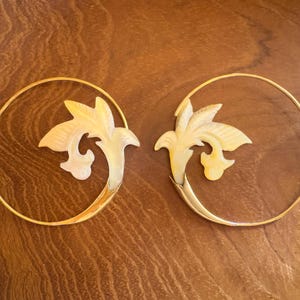 Peut inclure: Une paire de boucles d'oreilles créoles dorées, chacune ornée d'un motif floral stylisé de couleur crème. Les boucles d'oreilles sont posées sur un fond en bois chaleureux, mettant en valeur leur esthétique élégante et naturelle.