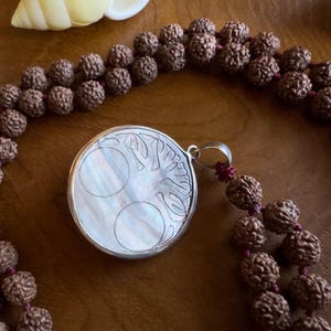 May include: A brown beaded mala necklace with a silver pendant. The pendant features a tree of life design with a mother-of-pearl inlay. The beads are dark brown, and the necklace rests on a wooden surface.