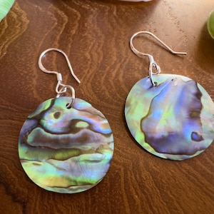 Peut inclure: Une paire de boucles d'oreilles en coquillage d'ormeau irisé avec des crochets argentés. Les boucles d'oreilles sont rondes et plates, affichant des teintes bleues, vertes et violettes. Elles sont posées sur une surface en bois brun foncé.