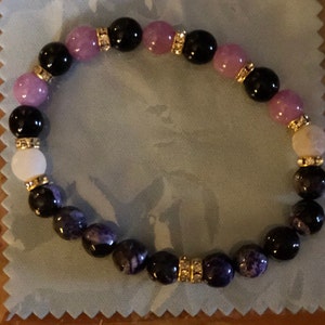 May include: A purple, black, and white beaded bracelet with gold accents. The bracelet is made with round beads and has a stretchy band.