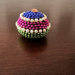 May include: A small, round, decorative box made of beads. The box is layered with green, pink, white, and blue beads. The top of the box has a gold accent.