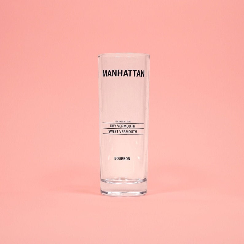 May include: A clear, rectangular Manhattan cocktail glass with black text. The text includes "MANHATTAN", ingredient instructions, and "BOURBON". The glass is set against a solid pink background.