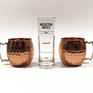 May include: Two copper Moscow Mule mugs with hammered texture and brass handles flank a tall, clear Moscow Mule mixing glass. The glass has black text that reads "Moscow Mule" and ingredient lines for lime, ginger beer, and vodka.