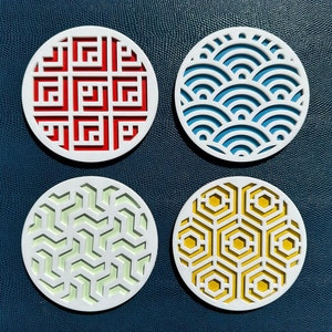 May include: Four white coasters with different geometric patterns. The coasters feature a red and white geometric pattern, a blue and white wave pattern, a green and white geometric pattern, and a yellow and white honeycomb pattern.