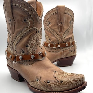 May include: A pair of tan leather cowboy boots with intricate stitching and stud details. The boots feature a pointed toe, a brown leather strap with silver embellishments, and a stacked heel. The boots are designed with a classic western aesthetic.