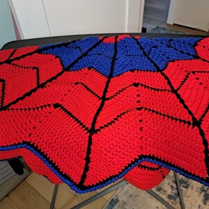 Puede incluir: Una manta de ganchillo hecha a mano con un dise&ntilde;o geom&eacute;trico en rojo y azul. La manta tiene un patr&oacute;n de estrella con l&iacute;neas negras que delinean las formas. Los bordes est&aacute;n acabados con un borde azul, creando un contraste llamativo.