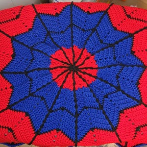 Puede incluir: Una manta de ganchillo hecha a mano en una vibrante combinaci&oacute;n de colores rojo, azul y negro. La manta presenta un dise&ntilde;o circular central con patrones radiantes, creando un efecto de explosi&oacute;n estelar. La manta se utiliza probablemente para la decoraci&oacute;n del hogar.