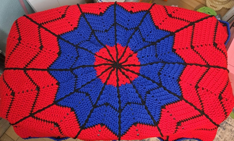 Puede incluir: Una manta de ganchillo hecha a mano en una vibrante combinaci&oacute;n de colores rojo, azul y negro. La manta presenta un dise&ntilde;o circular central con patrones radiantes, creando un efecto de explosi&oacute;n estelar. La manta se utiliza probablemente para la decoraci&oacute;n del hogar.