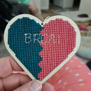 May include: A wooden heart-shaped ornament with a teal and pink cross-stitch design. The word "BRIA" is stitched in white thread on the ornament.