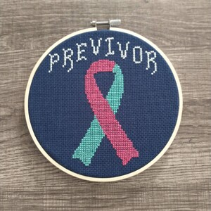 May include: A blue embroidery hoop with a pink and teal ribbon and the word "PREVIVOR" stitched in white.