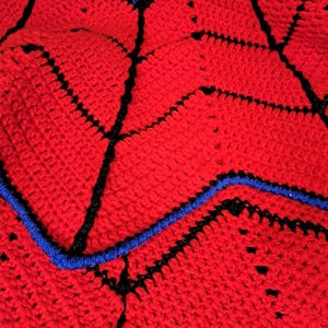 Puede incluir: Primer plano de una manta de ganchillo hecha a mano. La manta presenta una base roja vibrante con detalles en negro y azul. El dise&ntilde;o incluye l&iacute;neas negras y bordes azules, creando un patr&oacute;n geom&eacute;trico. La textura del ganchillo es visible.