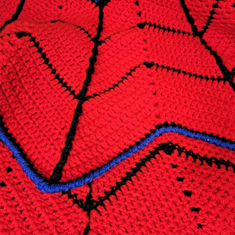 Puede incluir: Primer plano de una manta de ganchillo hecha a mano. La manta presenta una base roja vibrante con detalles en negro y azul. El dise&ntilde;o incluye l&iacute;neas negras y bordes azules, creando un patr&oacute;n geom&eacute;trico. La textura del ganchillo es visible.