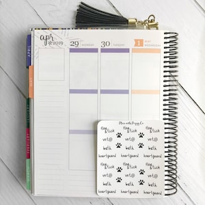 May include: A spiral-bound planner open to April 2019, with a black tassel and a sticker sheet. The sticker sheet has text including "flea & tick", "vet@", "bath", and "heartguard", along with paw print graphics. The planner is on a white wooden surface.