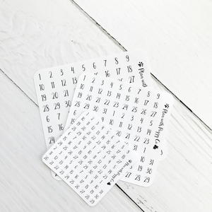 May include: White sticker sheets with black numbers, perfect for planning and organization. Each sheet features a different arrangement of numbers, with the text "Plan with Peggy Co" and a paw print logo.