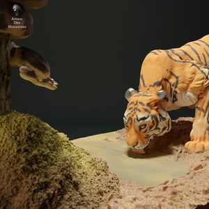 May include: A detailed miniature diorama featuring a tiger drinking water from a small pool. The tiger has orange fur with black stripes. A snake is coiled around a tree in the background. The diorama is set on a sandy base.